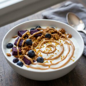 Yogurt Glow Bowl Topped with Purple Sweet Potato, Blueberries, Granola, Bee Pollen and Peanut Butter Drizzle